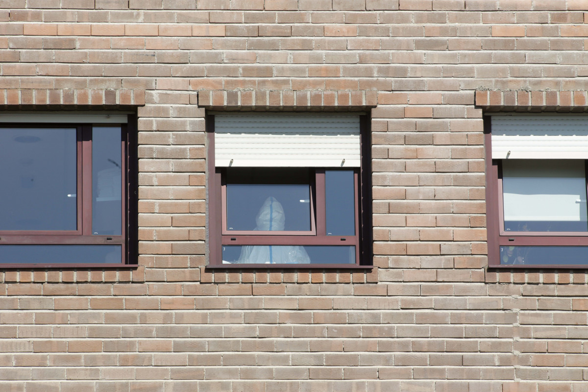 Archivo - Un trabajador sanitario con un EPI, en la ventana de una habitación de una residencia