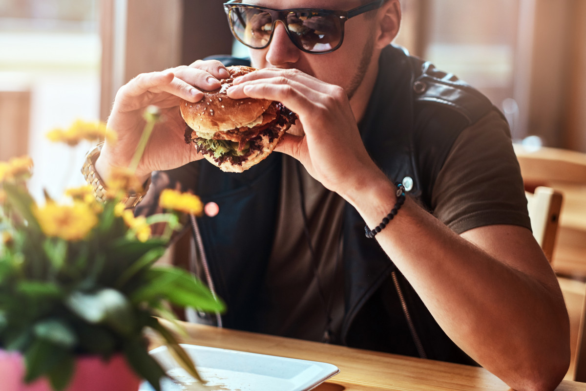 Hipster con un elegante corte de pelo y barba se sienta en una mesa decidio cenar en un cafe al borde de la carretera comiendo una hamburguesa
