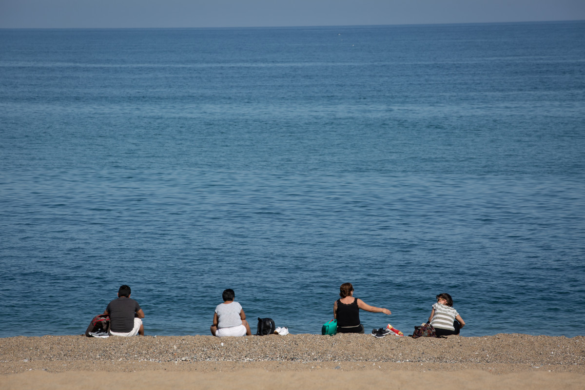 Diverses persones assegudes a la Platja de la Barceloneta respecten la distància de seguretat.
