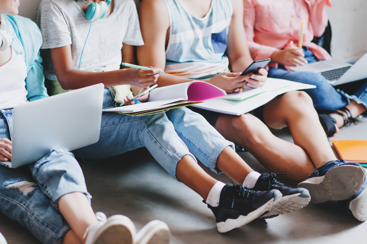 Noia jeans sabatilles negres escrivint conferència gran llibre text asseguda sòl amics universitat jove escrivint missatge telèfon mentre altres estudiants treballen ordinadors portàtils
